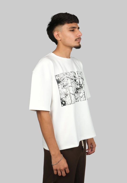 A man wearing a white oversized t-shirt with a black graphic print on the front, paired with dark brown pants, photographed against a solid white background.