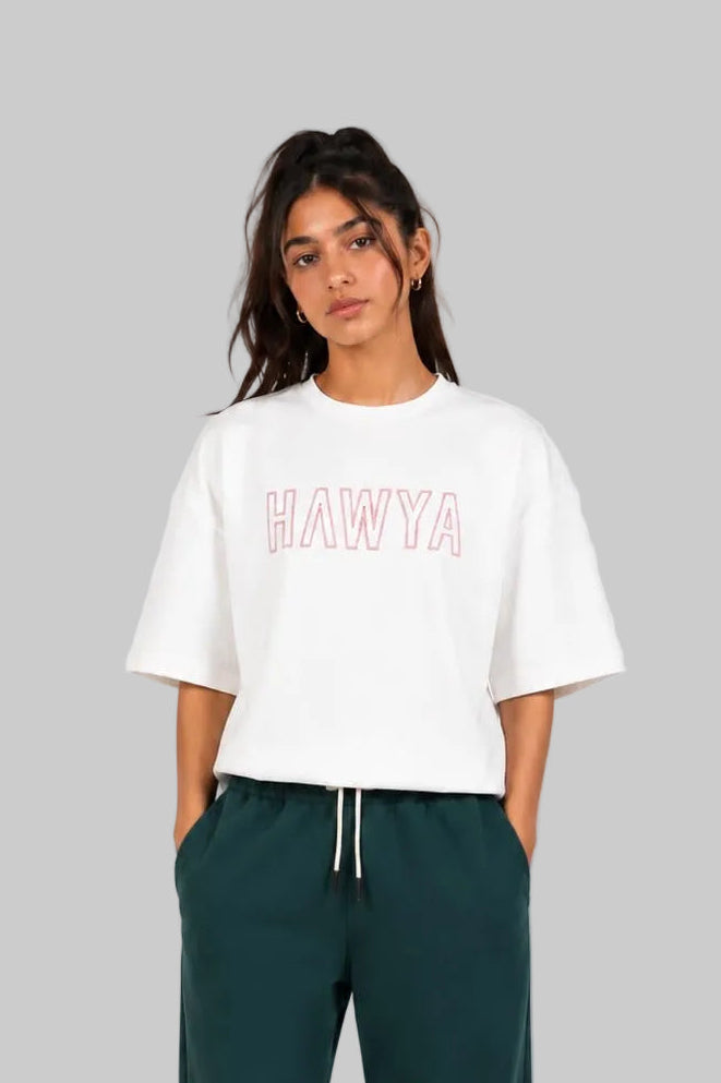 A model stands against a light gray studio background wearing a white cropped T-shirt with 'HAWYA' printed in pink outline letters across the chest, paired with dark green sweatpants and white sneakers.