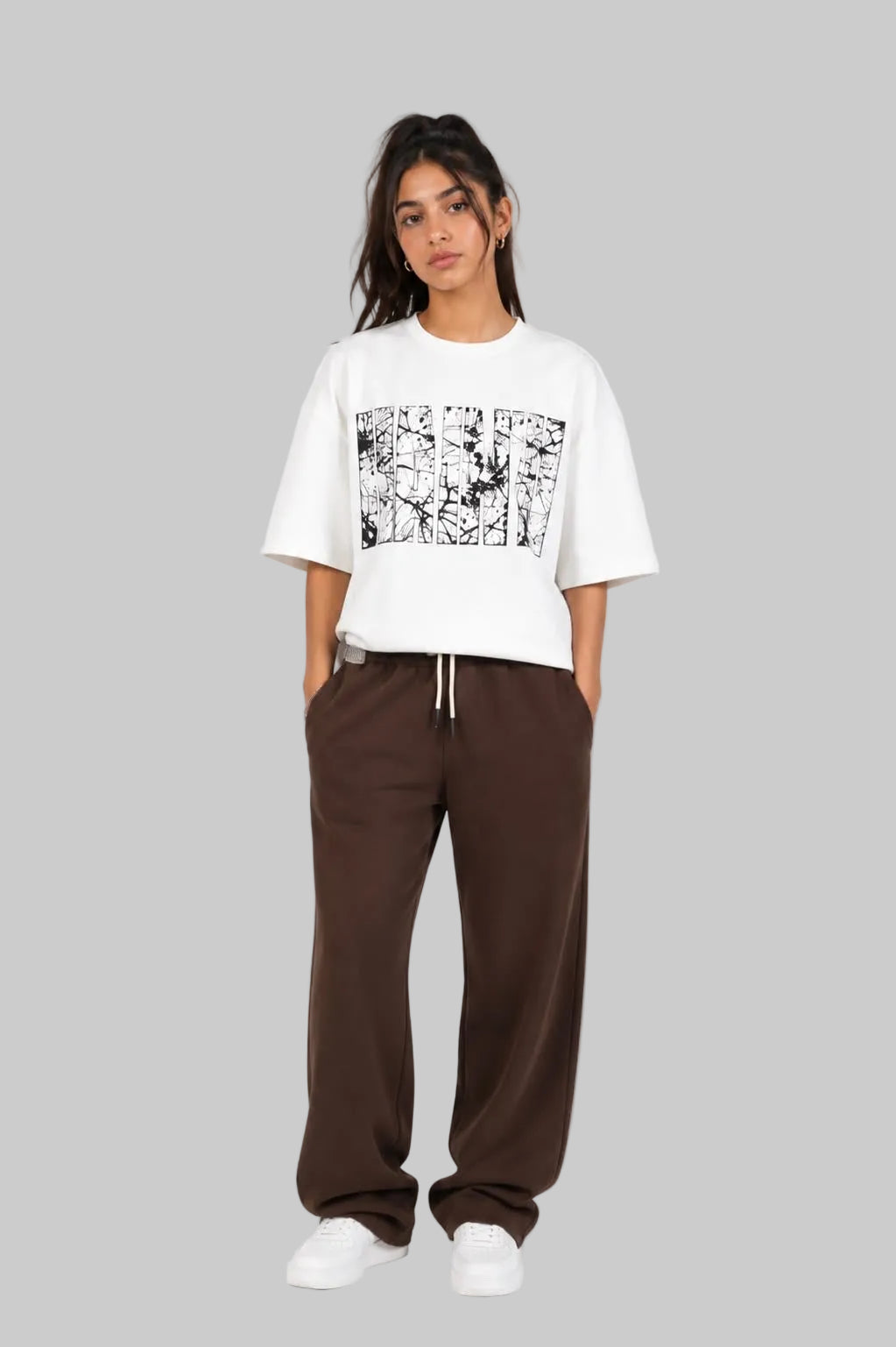 A model stands against a light gray studio background wearing a white cropped T-shirt with a black graphic print on the front, paired with dark brown wide-leg sweatpants and white sneakers.