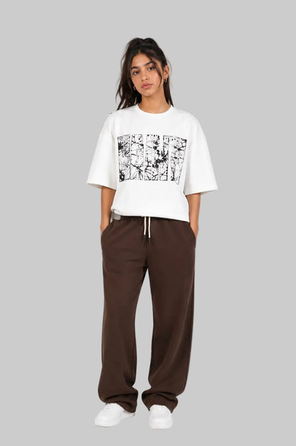 A model stands against a light gray studio background wearing a white cropped T-shirt with a black graphic print on the front, paired with dark brown wide-leg sweatpants and white sneakers.
