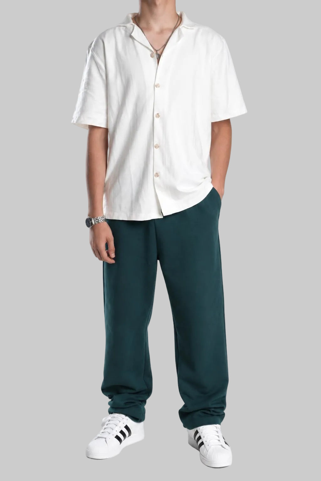 A model wearing a white short-sleeve button-up shirt, dark green relaxed-fit pants, and white sneakers with black stripes, standing against a plain white studio background.