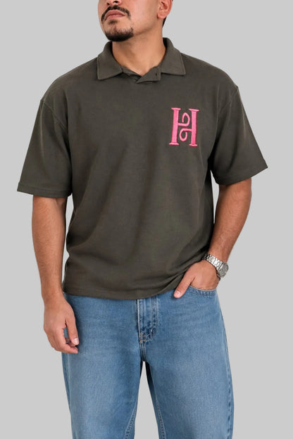 A man wearing a dark olive green short-sleeve polo shirt with a pink embroidered 'H' logo on the left chest, paired with blue jeans, against a plain light gray background.