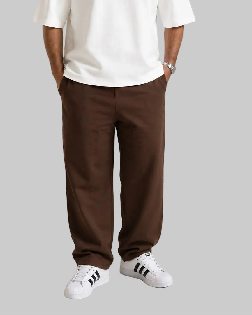 A man wearing a white oversized t-shirt with 'HAWYA' printed in pink outline text on the chest, paired with brown pants and white sneakers, standing in a studio setting next to a wooden bench.