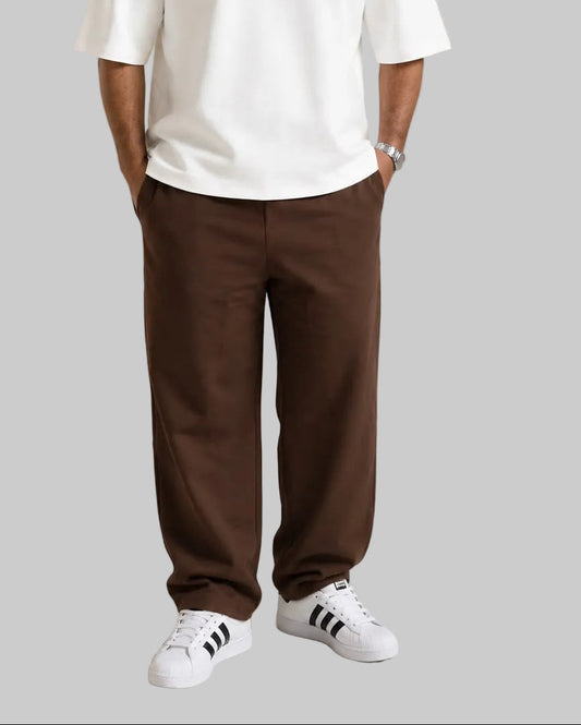 A man wearing a white oversized t-shirt with 'HAWYA' printed in pink outline text on the chest, paired with brown pants and white sneakers, standing in a studio setting next to a wooden bench.