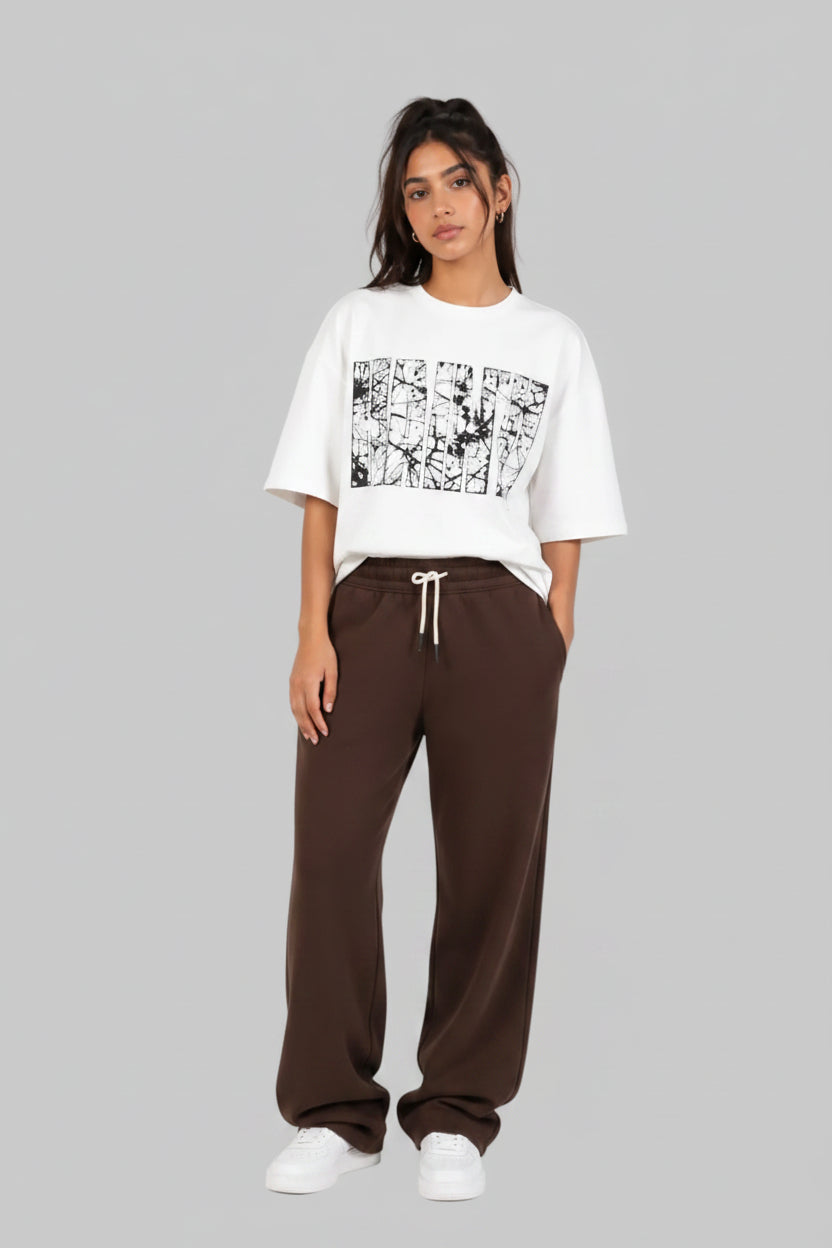 A model stands against a light gray studio background wearing a white cropped T-shirt with a black graphic print on the front, paired with dark brown wide-leg sweatpants and white sneakers.
